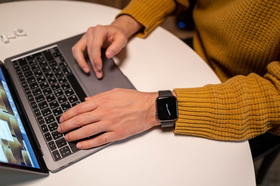 Adult wearing a smartwatch, typing on a laptop at a table. Wireless earbuds nearby.