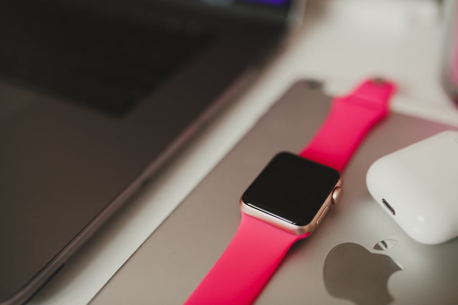 A stylish smartwatch with a pink band placed on a laptop surface, surrounded by modern gadgets.