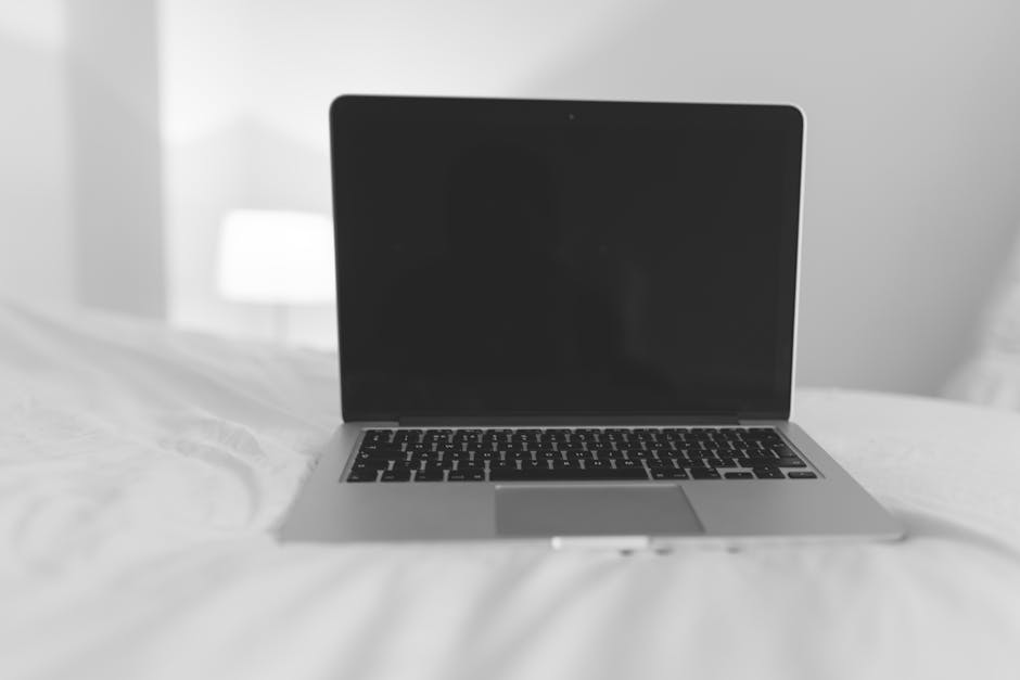 A laptop sits on a bed in a minimalist white room, perfect for remote work inspiration.