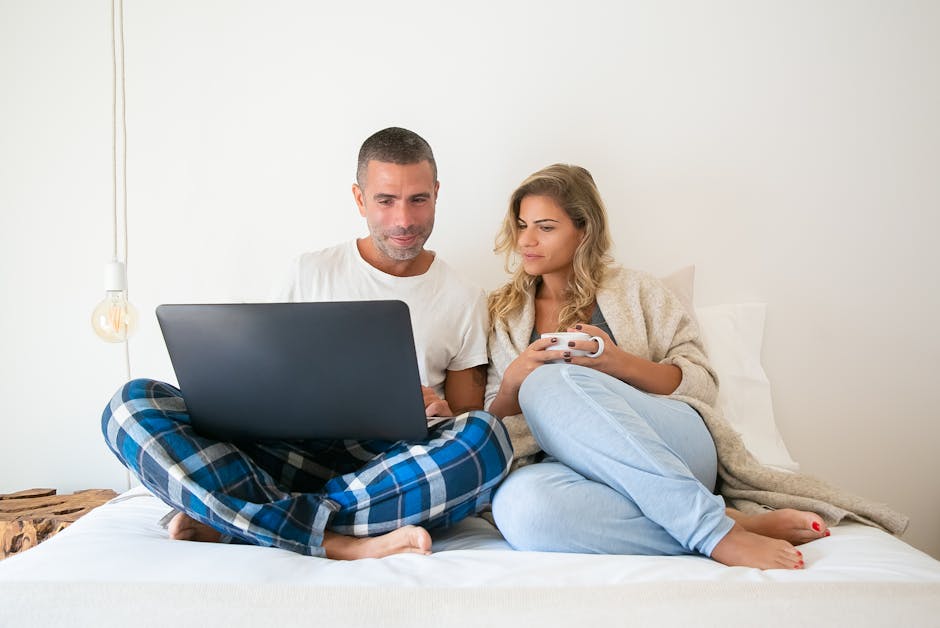 A couple in casual wear working remotely from bed, sharing a laptop in a cozy home setting.