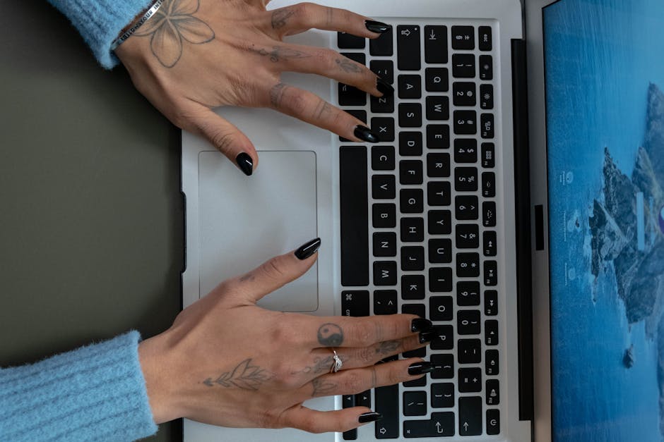 Close-up of tattooed hands with black nails typing on a laptop keyboard indoors.