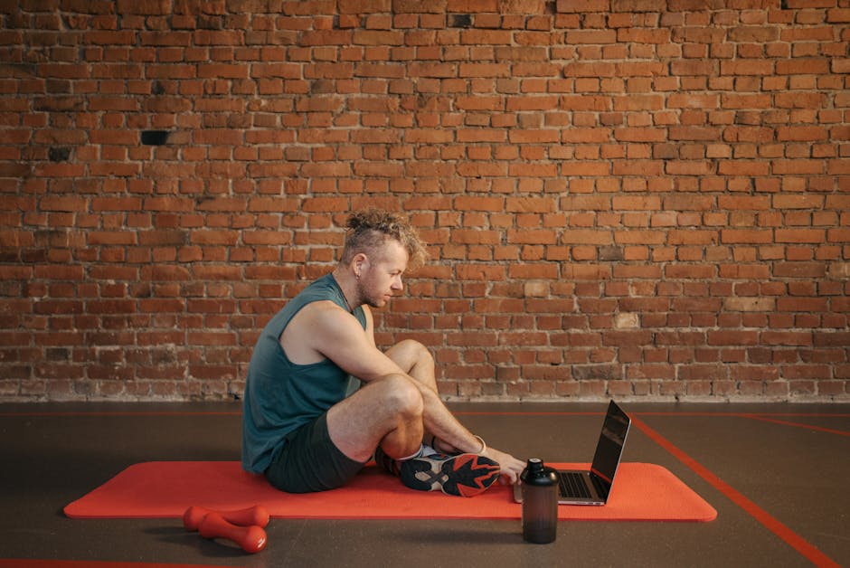 A man sits on a yoga mat using a laptop, emphasizing home fitness and technology.