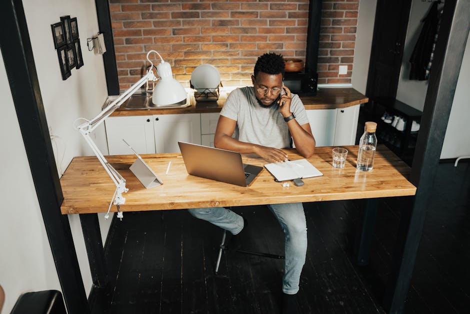 A young man at a desk in a home office, using a laptop and phone, embodying remote work lifestyle.