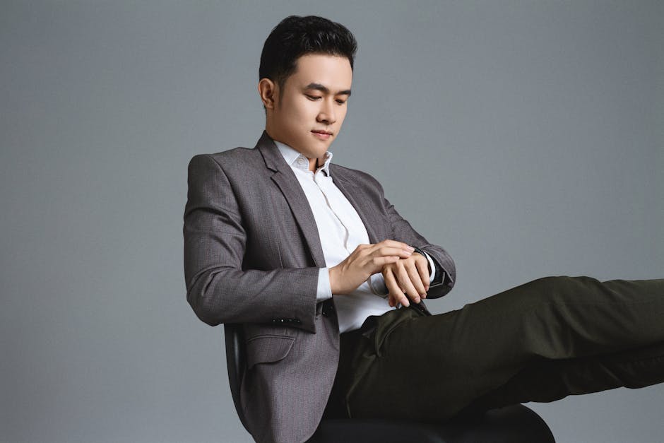 Asian businessman in a stylish suit looking at his watch, sitting in a studio setting.