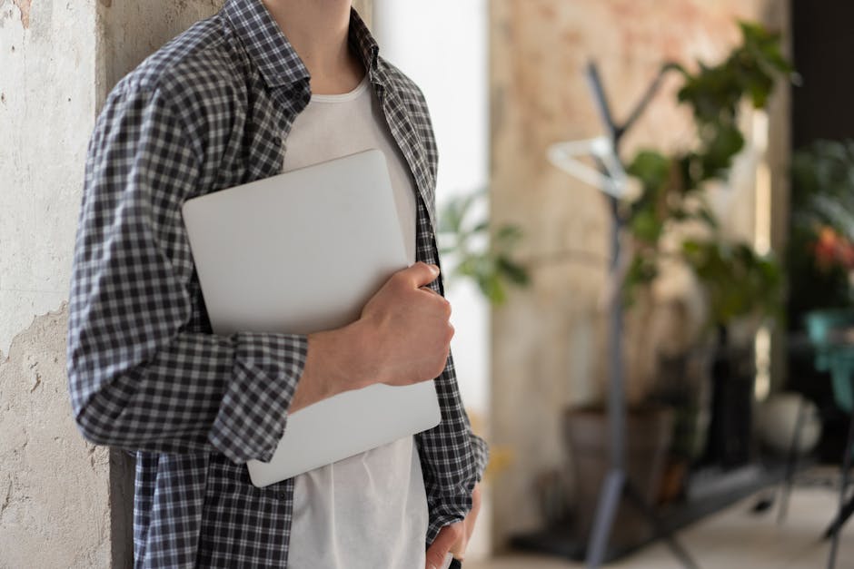 Young adult in casual attire holding laptop in bright indoor setting.