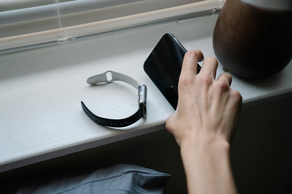 Close-up of a hand holding a smartphone next to a smart watch on a windowsill, indoors.