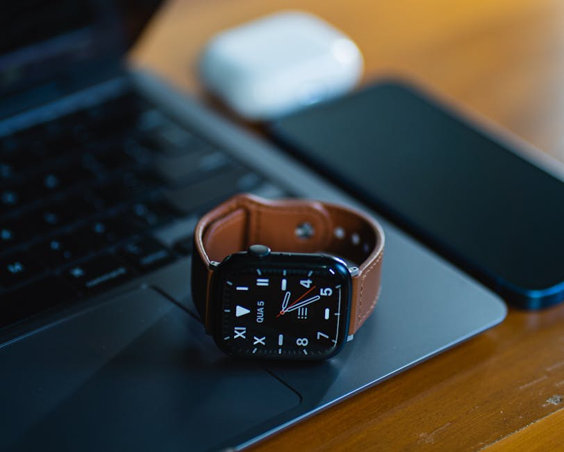 A smartwatch resting on a laptop keyboard beside a smartphone and earbuds.
