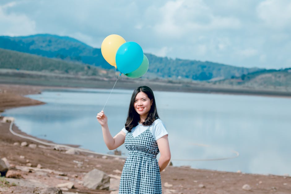 A vibrant woman enjoys a sunny day, holding balloons by a tranquil lake.