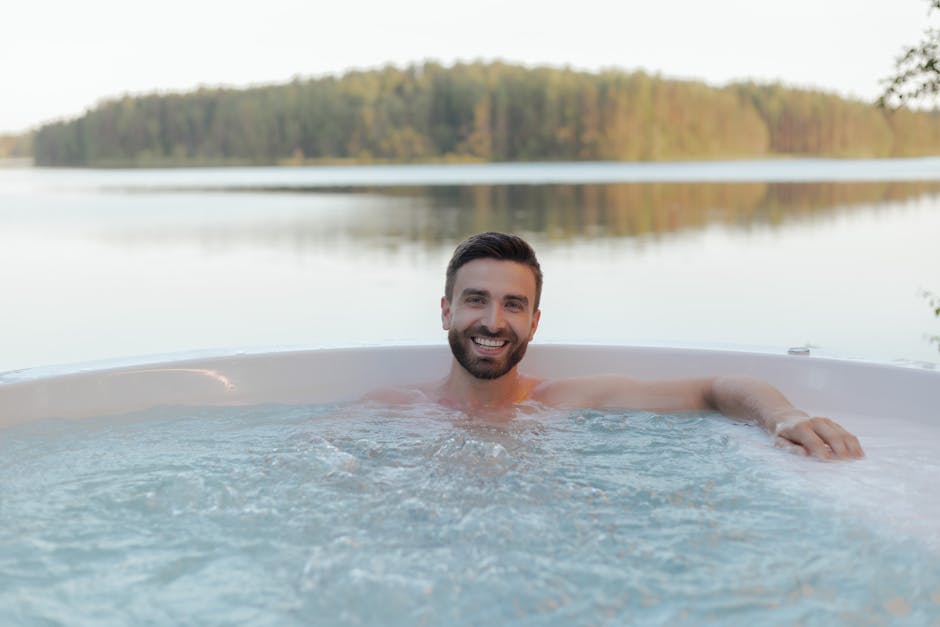 Shirtless man smiling in an outdoor hot tub by a serene lake, enjoying relaxation and wellness.